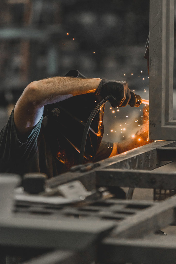 Man wearing protective welding gear as he uses welding equipment, partially blocked by metal and steel bars in foreground of photo