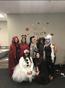 7 women in halloween costumes standing in front of office wall with the words Trick or Treat