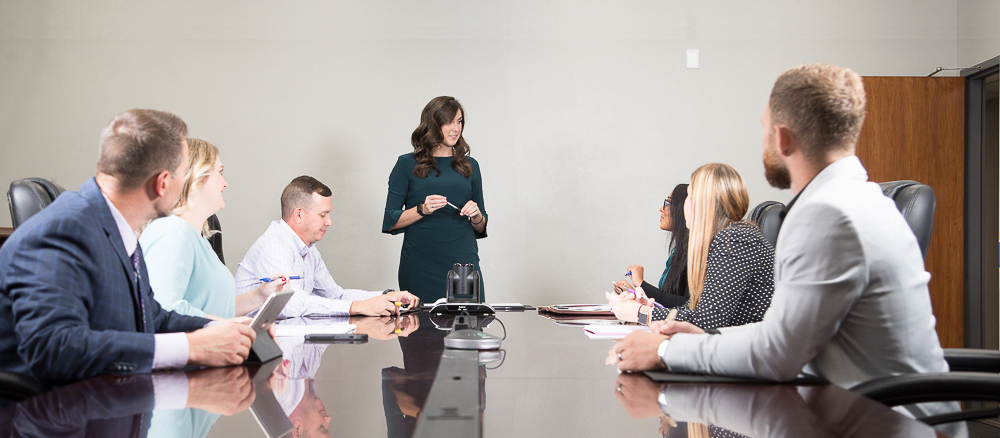 Woman in green business dress standing and talking to 6 people in business attire who are seated at a conference table.